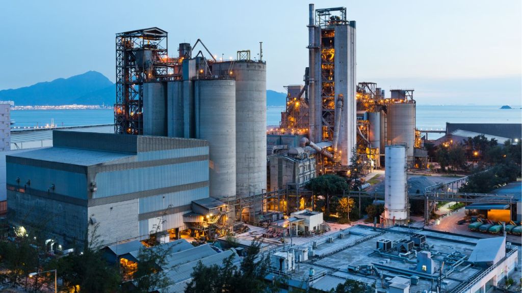 A cement manufacturing plant with large silos and industrial structures, set against a mountainous backdrop during twilight.