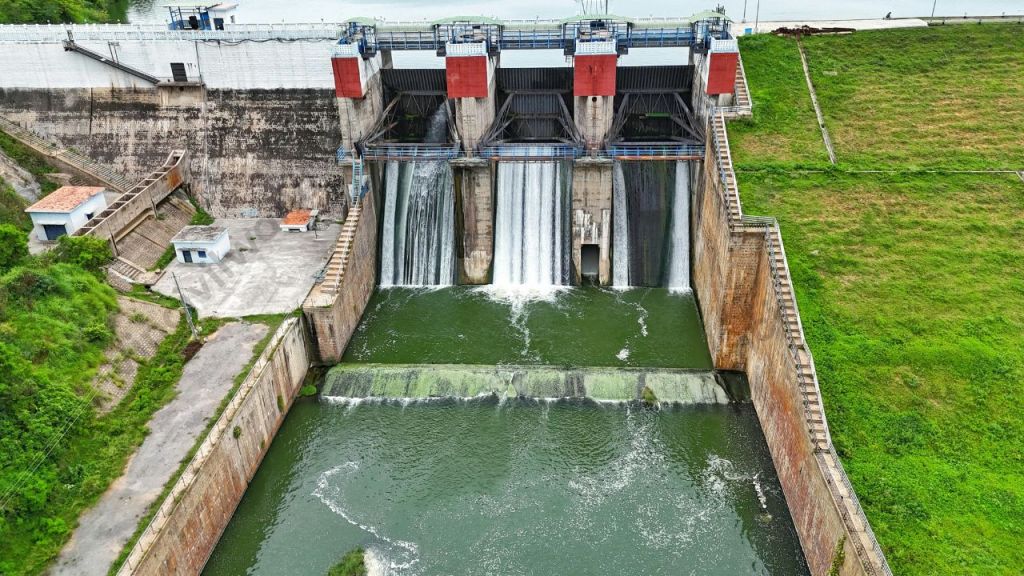 An aerial view of a dam structure with water flowing through gates, surrounded by green land.