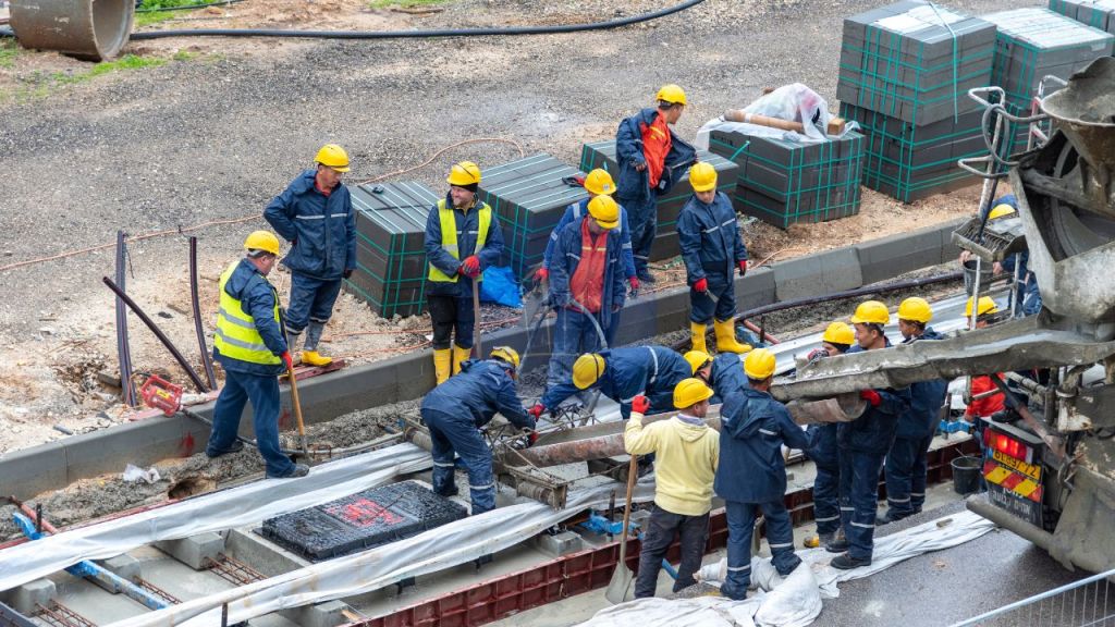 A group of construction workers in hard hats and safety gear, collaborating on a railway construction site, with stacks of materials and machinery visible in the background.