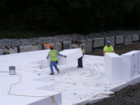 Construction workers handling large blocks of geofoam material on a site, with gravel and concrete barriers in the background.