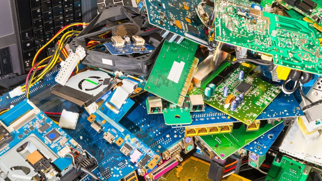 A cluttered pile of various electronic circuit boards and components scattered around, featuring an assortment of colors and types, with a keyboard and wires visible in the background.