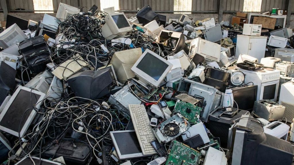 A large pile of discarded electronic devices, including old computers, monitors, keyboards, and tangled cables, in a warehouse setting.