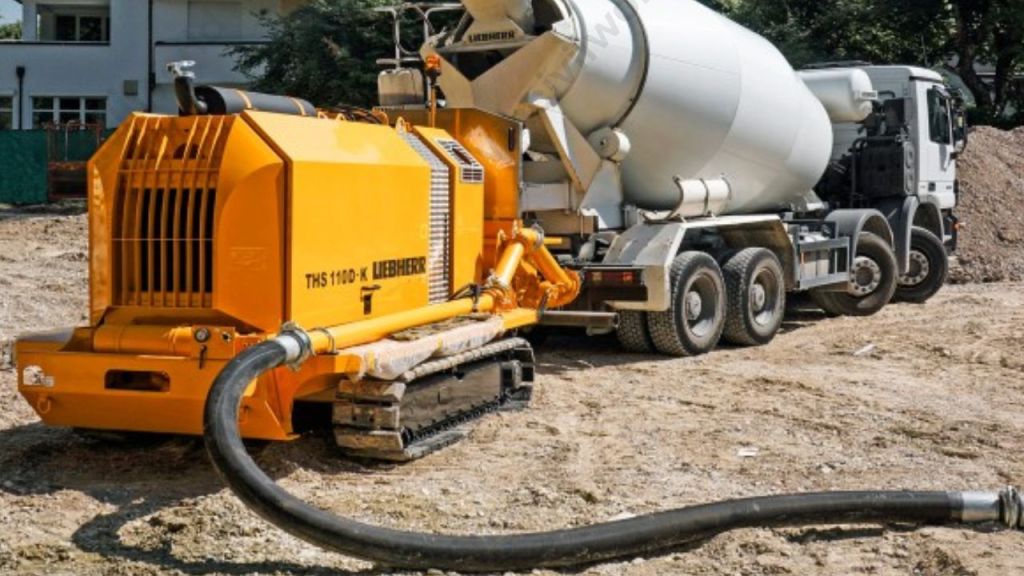 A stationary concrete pump connected to a cement mixer truck, with flexible hoses on the ground for concrete delivery at a construction site.