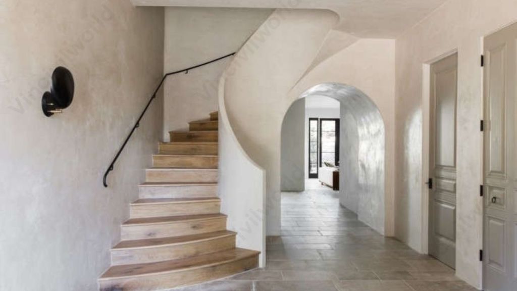 An interior view of a modern home featuring a wooden staircase, smooth white plastered walls, and tiled flooring. The design includes a curved wall that leads to an arched entrance.