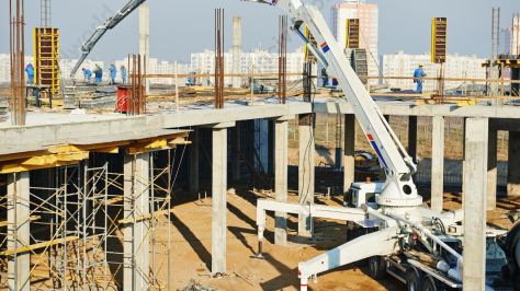 A truck-mounted concrete pump with a long boom is positioned on a construction site, delivering concrete from a mixer to the elevated framework of a building under construction, surrounded by workers and scaffolding.