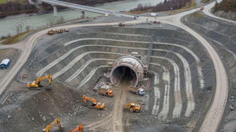 Aerial view of a tunnel construction site, showing excavation work and heavy machinery around a partially completed tunnel entrance.
