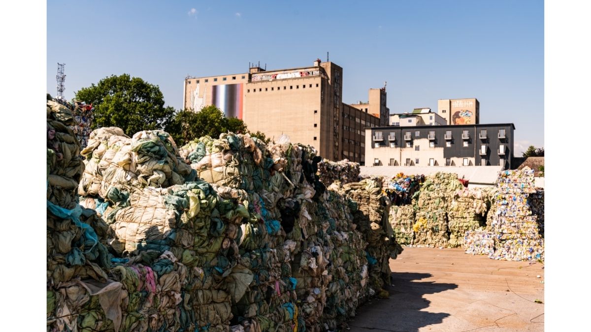 A large pile of compressed plastic waste and discarded materials in an industrial area, with buildings and greenery in the background.
