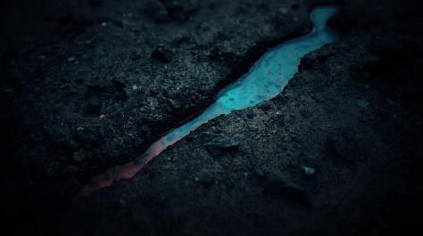 A close-up view of a crack in the ground, through which bright blue liquid is visible, contrasting against the dark soil.
