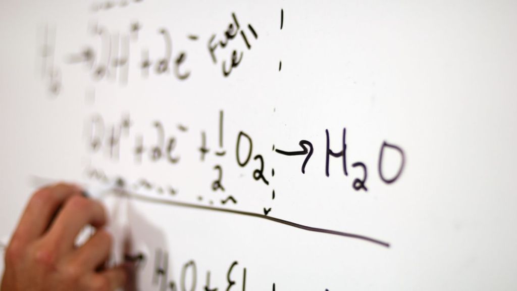 A person writing chemical equations related to fuel cell reactions on a whiteboard, demonstrating the transformation of hydrogen and oxygen into water.