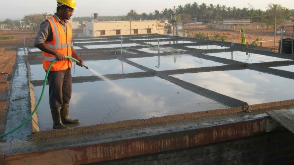 A construction worker applies water to a freshly poured concrete slab using a hose, surrounded by temporary containment for effective curing.