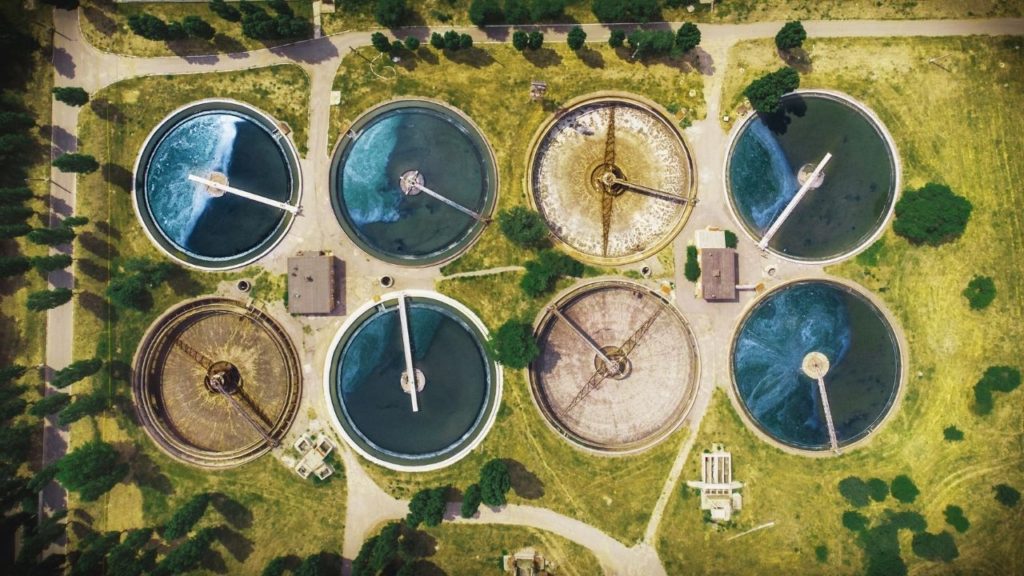 Aerial view of multiple circular wastewater treatment tanks with varying water clarity, surrounded by greenery.